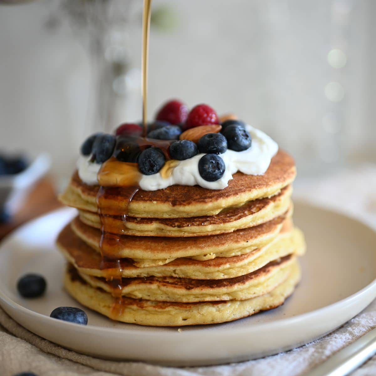 Stacked protein pancakes topped with whipped cream and berries and syrup being poured on them.