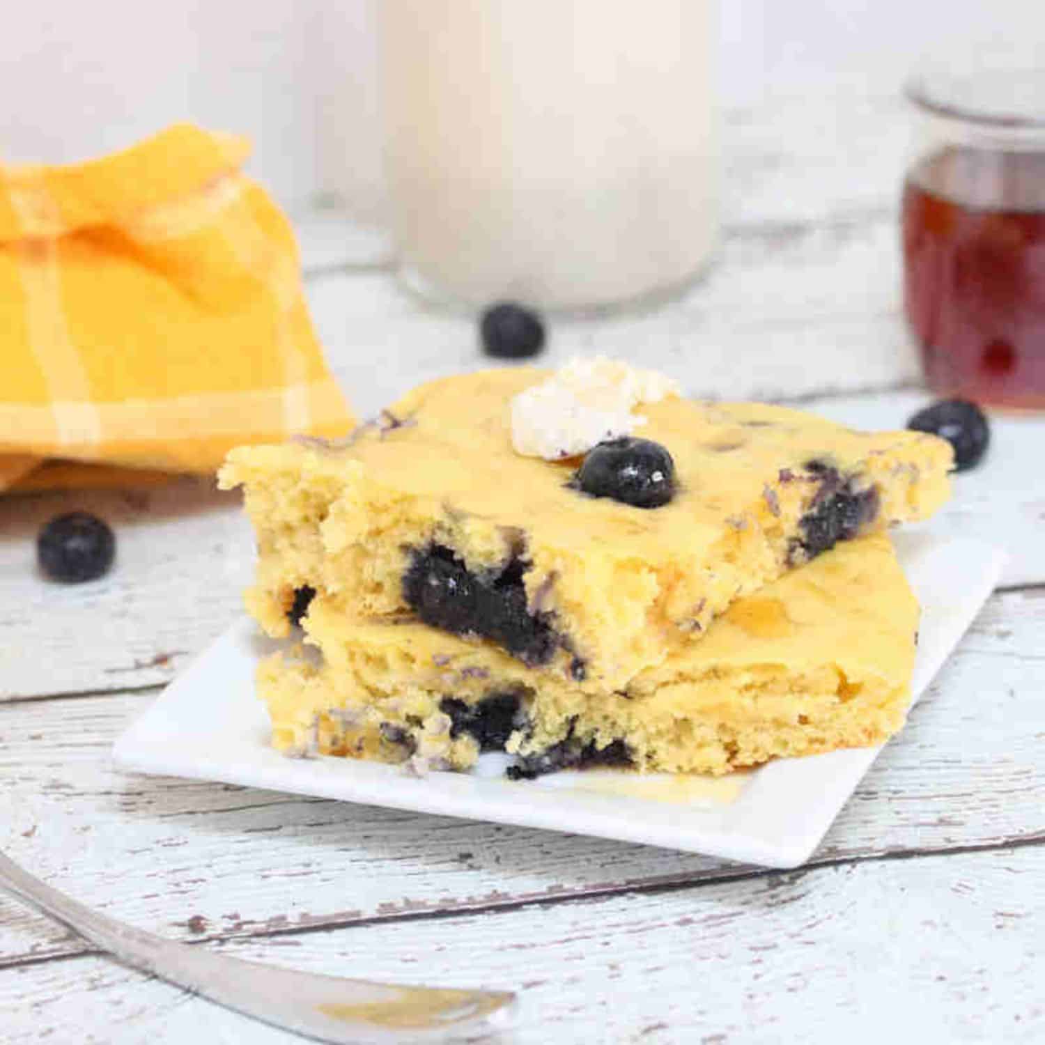 Protein pancakes with blueberries stacked on a white plate with maple syrup and milk behind them.