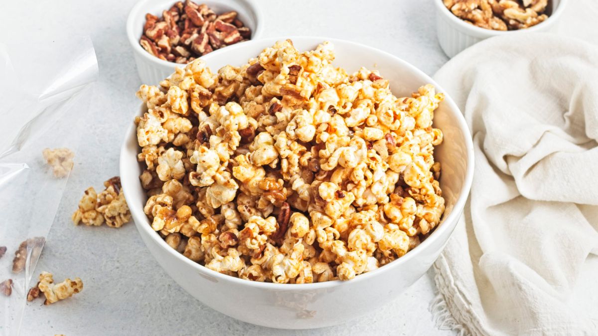 A white bowl filled with caramel popcorn and more popcorn around the bowl with chopped pecans behind it.