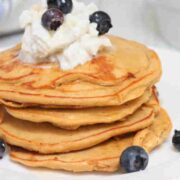 Protein pancakes stacked on a white plate topped with coconut whipped cream and blueberries.