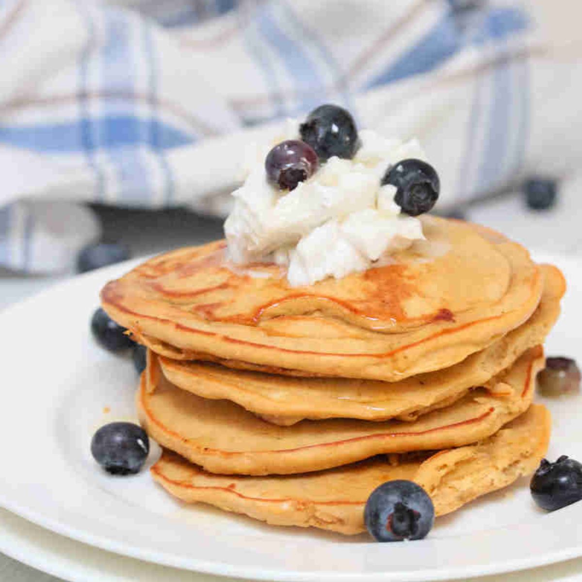 Protein pancakes stacked on a white plate topped with coconut whipped cream and blueberries.