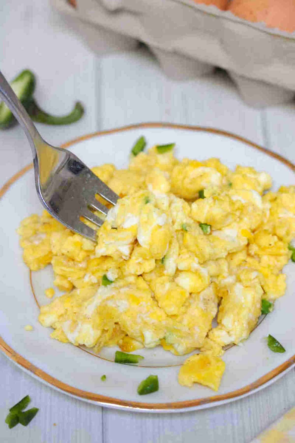 Scrambled eggs without milk on a white plate with a fork in them and topped with jalapenos.