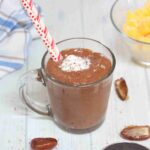 Chocolate smoothie with coconut milk on top and two red straws in a glass with dates around it.