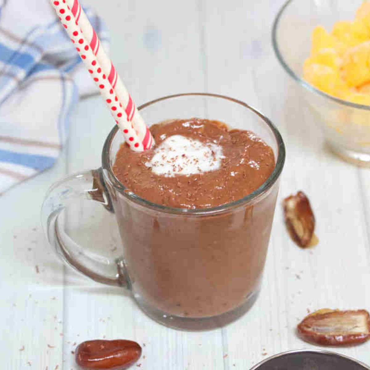 Chocolate smoothie with coconut milk on top and two red straws in a glass with dates around it.