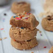 Three protein cookies stacked and topped with sprinkles the top cookie with a bite out of it and more cookies behind them.
