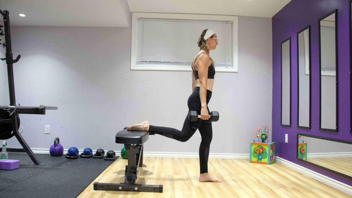 Woman in black workout clothes holding a dumbbell at her side with her back foot on a bench.