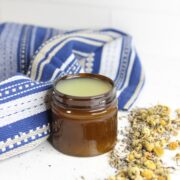 Amber container with homemade salve in it with a blue linen and chamomile flowers around it.
