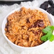 White bowl with shredded chipotle chicken in it and a slow cooker behind it.