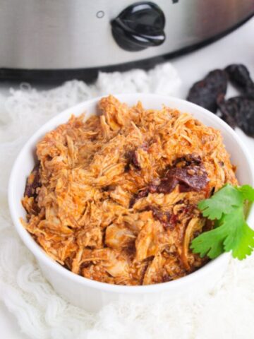 White bowl with shredded chipotle chicken in it and a slow cooker behind it.