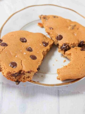 White plate with large single serving protein cookie broken in half with chocolate chips in it.