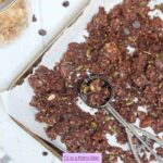 Chocolate granola on a baking tray with parchment paper with an ice cream scoop in it.