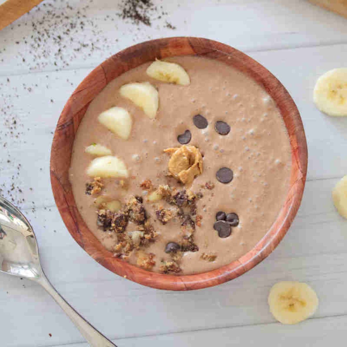 Wooden bowl with a coffee protein smoothie in it topped with granola, chocolate chips, bananas, and peanut butter.