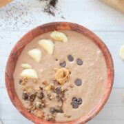 Wooden bowl with a coffee protein smoothie in it topped with granola, chocolate chips, bananas, and peanut butter.