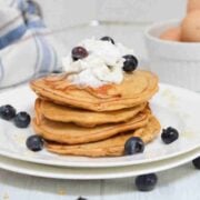 Four protein pancakes stacked on a white plate topped with coconut whipped cream and blueberries.
