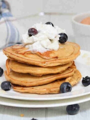 Four protein pancakes stacked on a white plate topped with coconut whipped cream and blueberries.