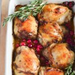 Baked chicken thighs in a white baking pan with cranberries and rosemary.