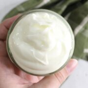 Small jar with whipped tallow lotion being held in a hand with a green linen behind it