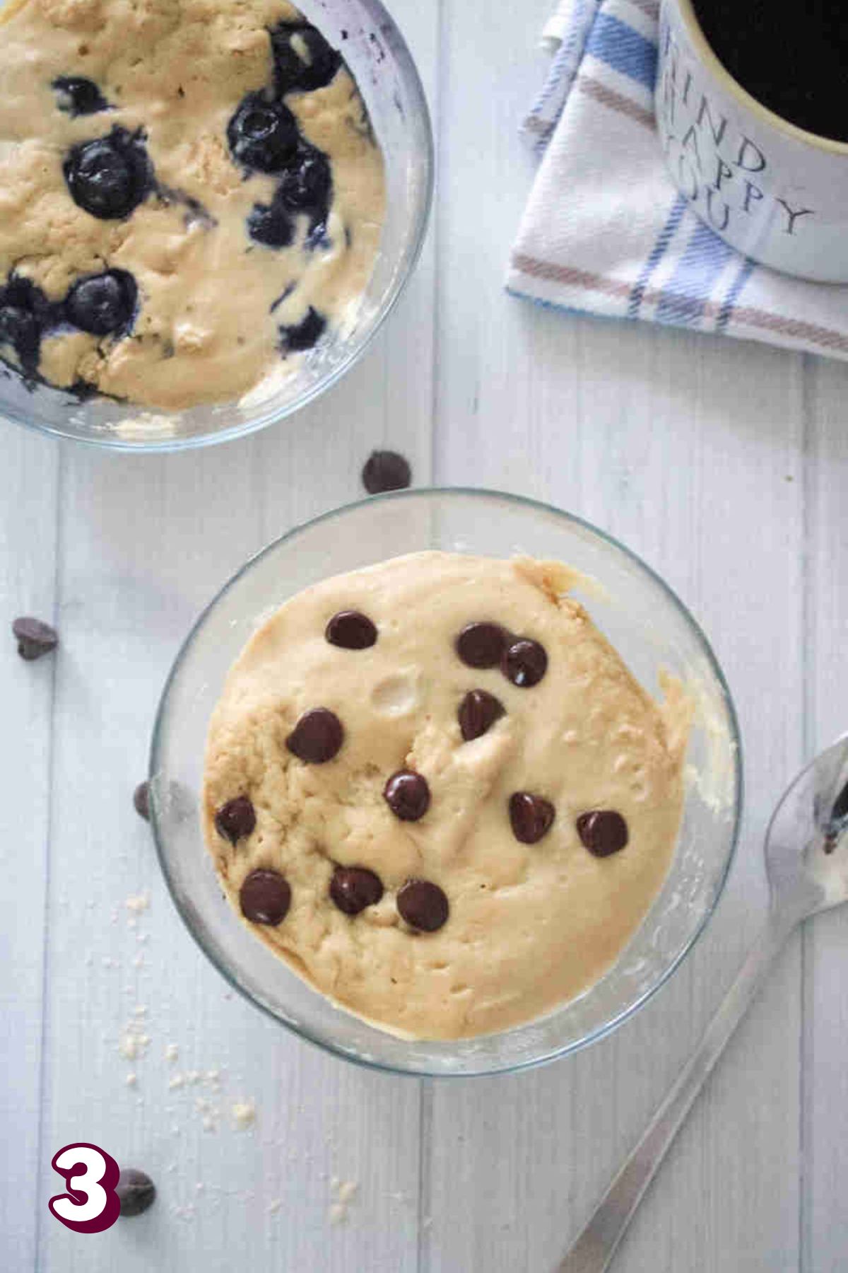 Two glass bowls with a protein mug cake in them one topped with chocolate chips and the other blurberries.