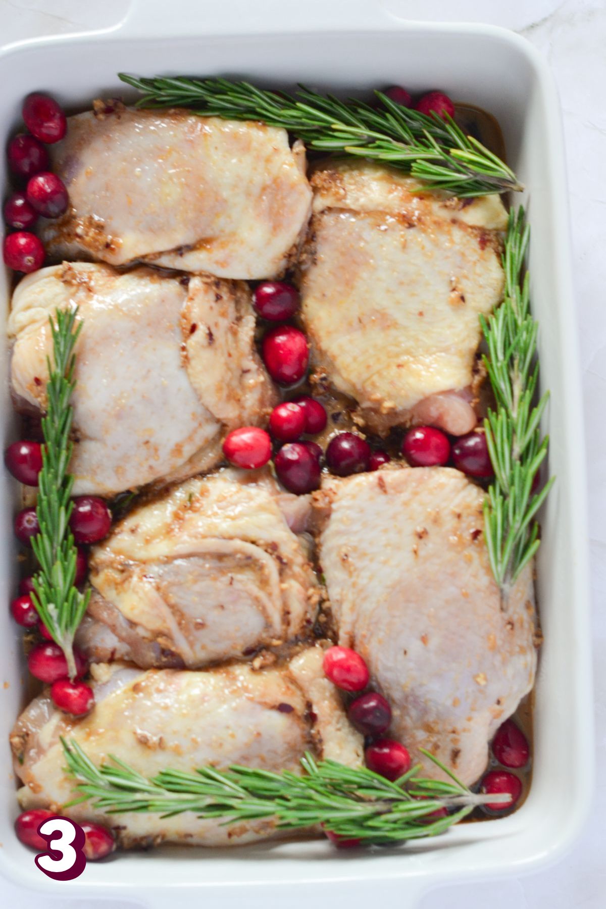 Raw chicken thighs in a white baking pan with cranberries and rosemary.
