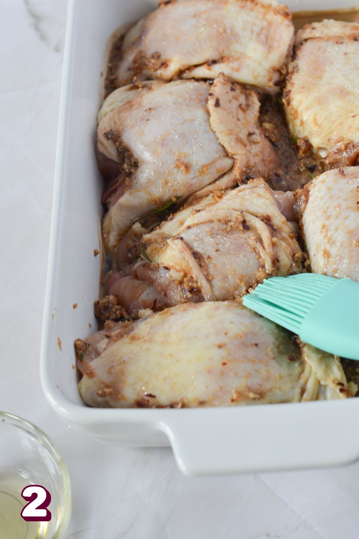 Blue brush brushing the tops of raw chicken thighs in a white baking dish.