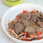 White bowl with gluten-free noodles, steak, leeks, and carrots with toppings behind it.