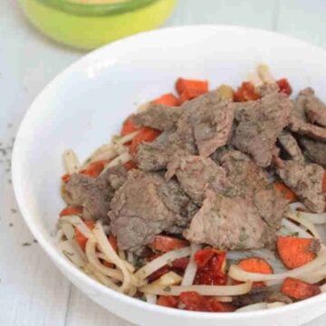 White bowl with gluten-free noodles, steak, leeks, and carrots with toppings behind it.