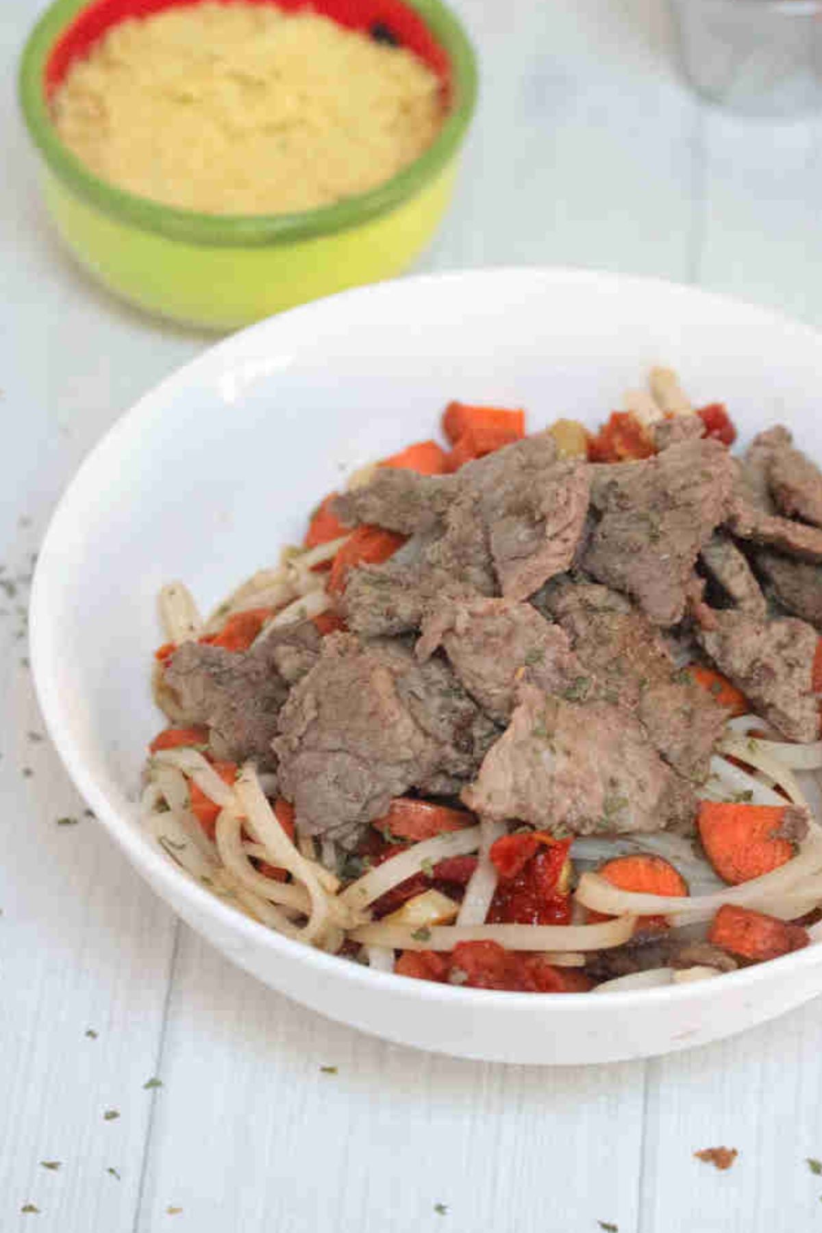 White bowl with gluten-free noodles, steak, leeks, and carrots with toppings behind it.