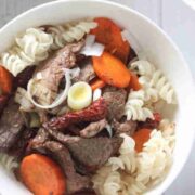White bowl with gluten-free noodles, steak, leeks, and carrots with more in a pan behind it.