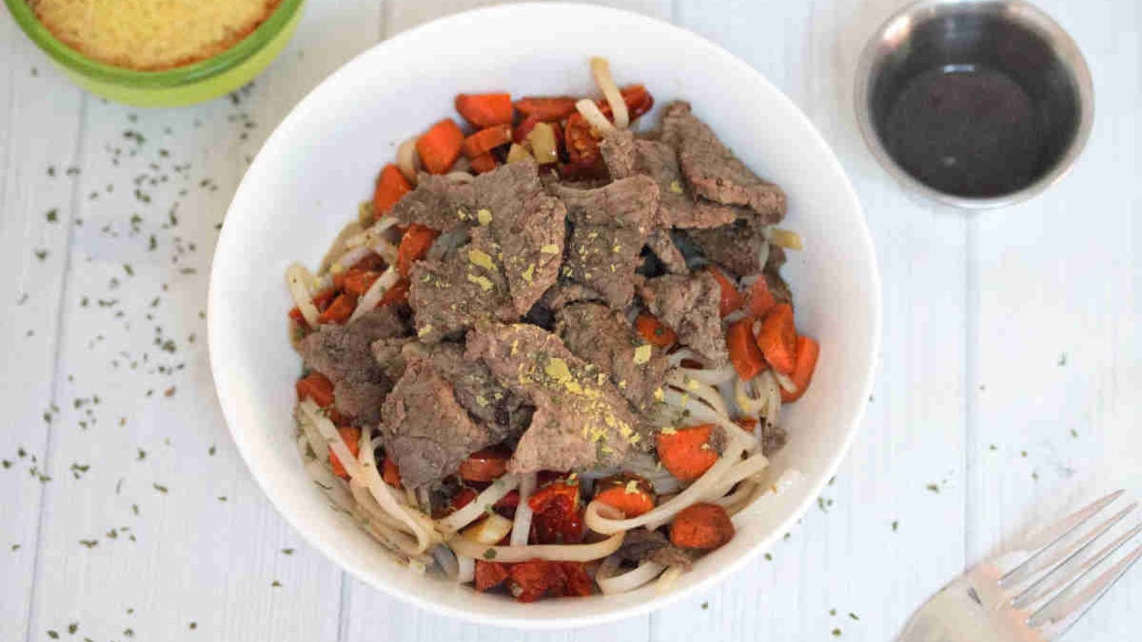 White bowl with gluten-free noodles, steak, leeks, and carrots with sauce and nutritional yeast behind it.