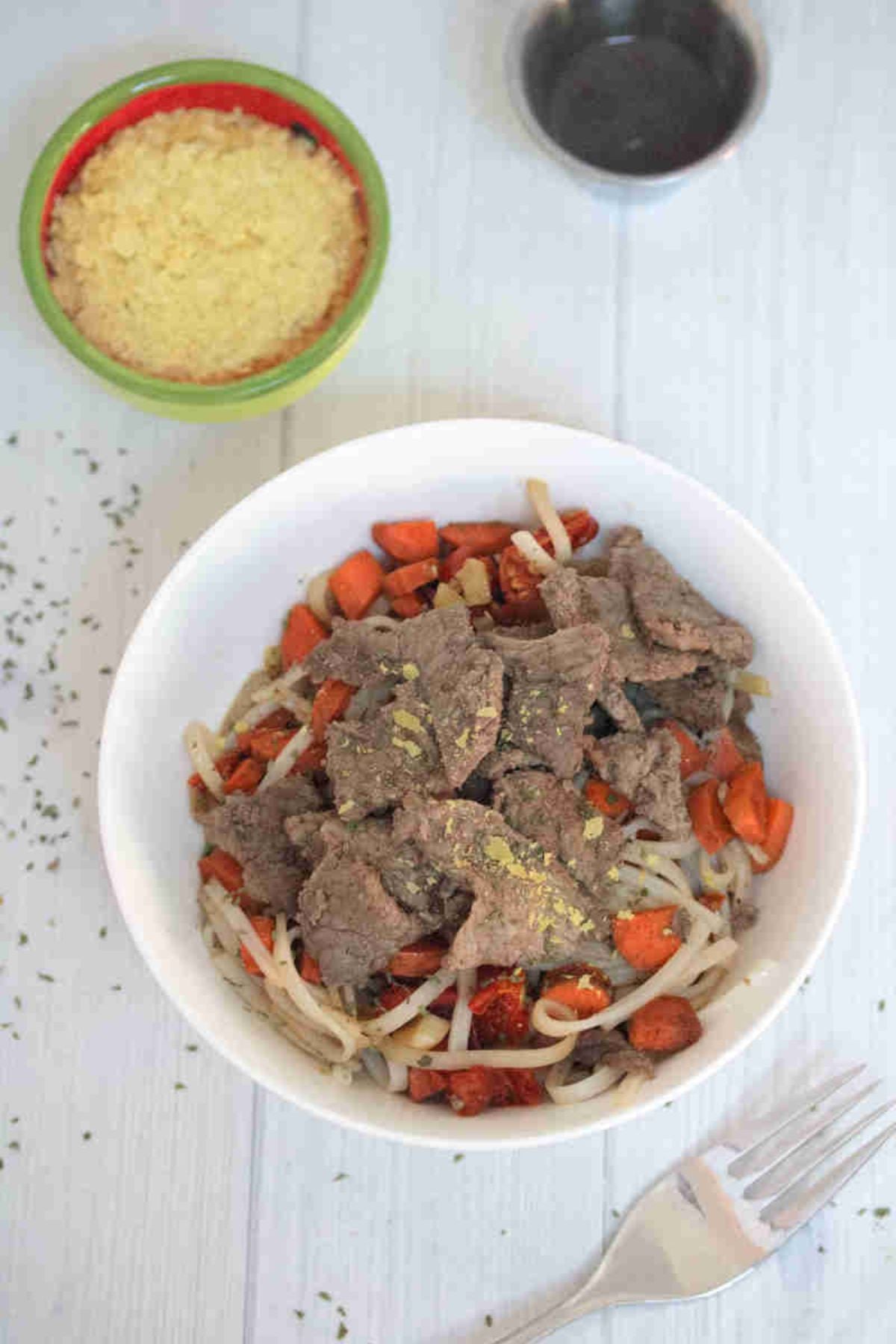 White bowl with gluten-free noodles, steak, leeks, and carrots with toppings behind it.