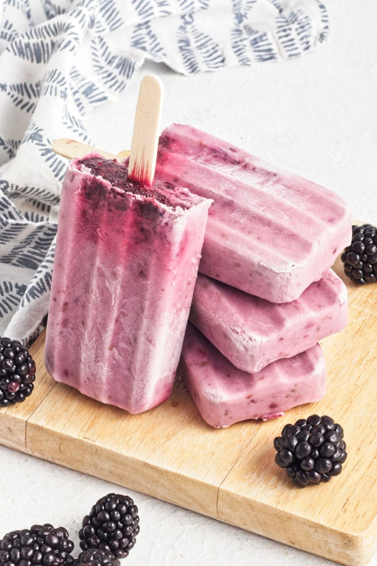 Black berry popsicles stacked on top of each other on a wooden board with blackberries around them.