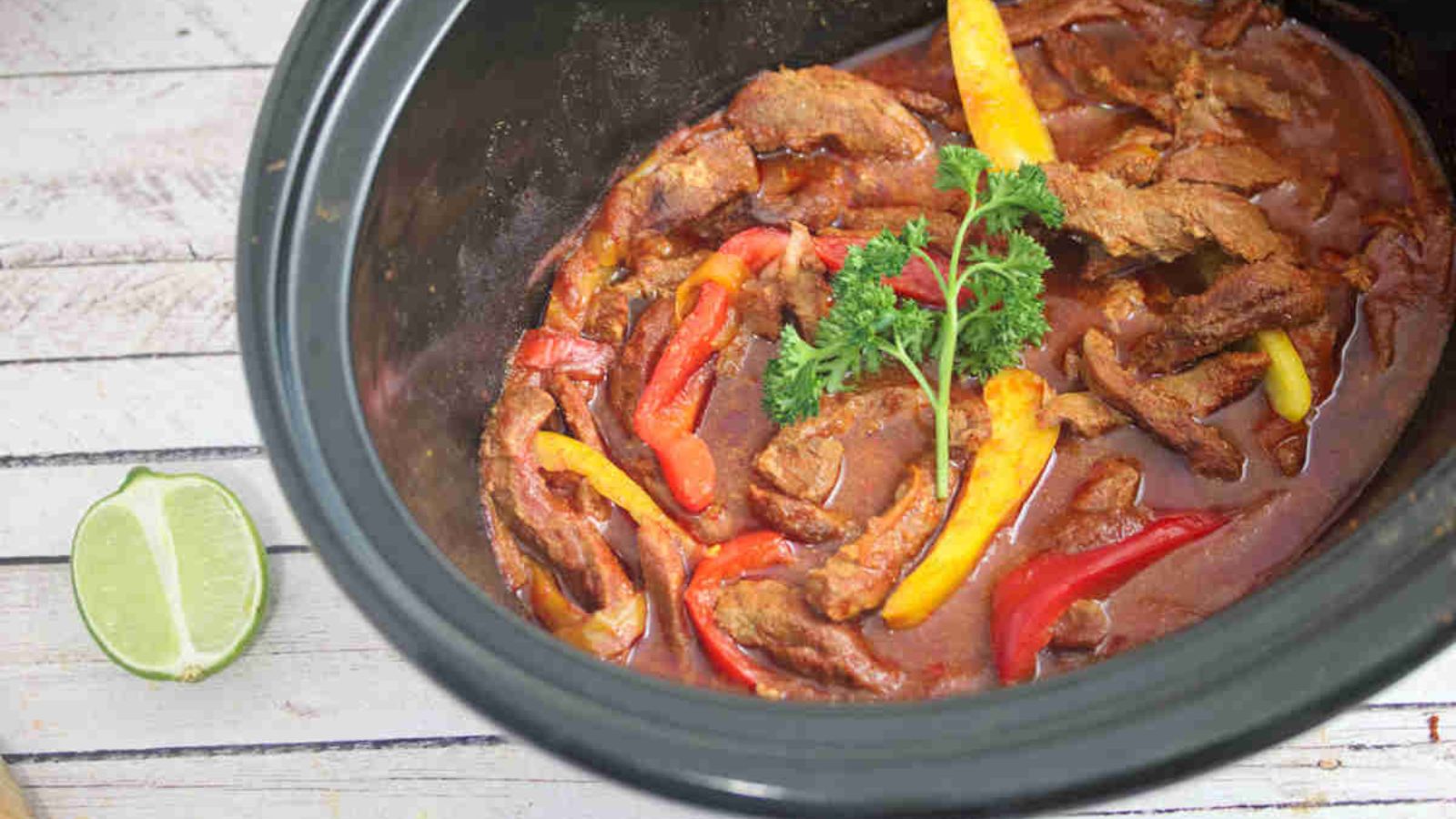 Beef fajitas in tomato sauce with peppers in a black slow cooker.