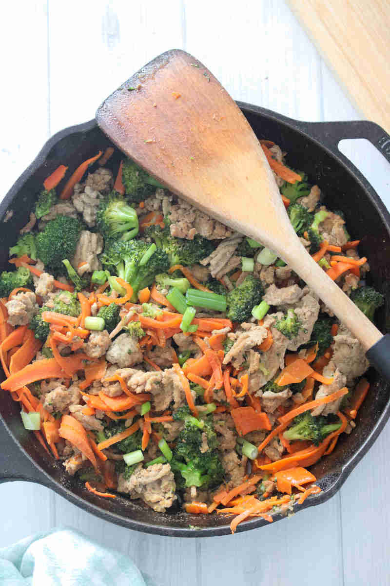 Ground turkey in a cast iron pan with carrots and broccoli with a wooden spoon on top.