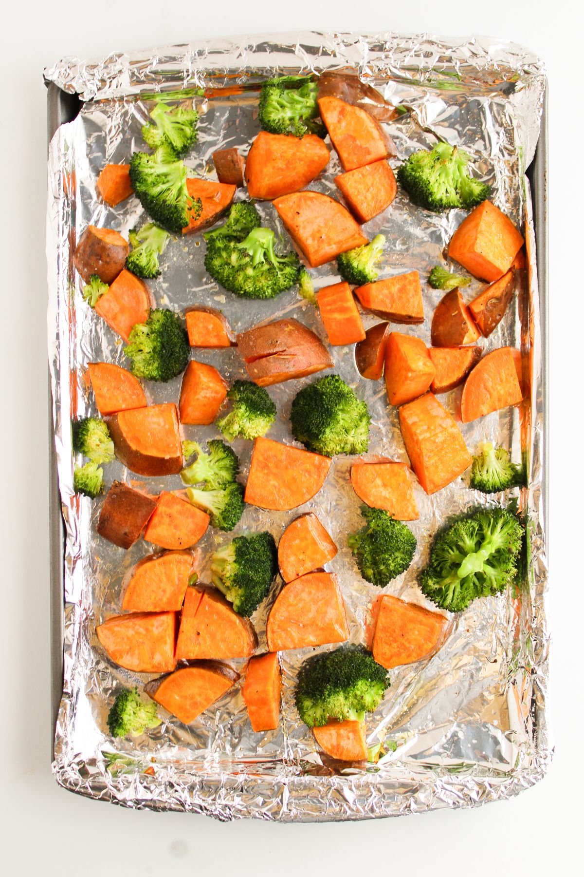 Sweet potatoes and broccoli tossed in oil and seasonings in a sheet pan with foil.
