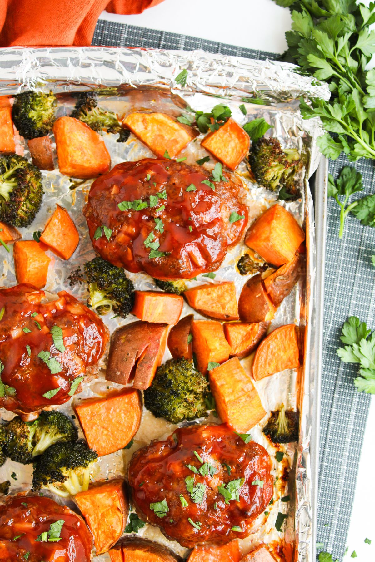 Ground beef meat loaves brushed with barbecue sauce with sweet potatoes and broccoli on a sheet pan with foil.