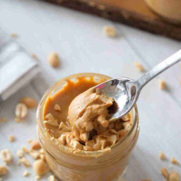 Homemade peanut butter in a mason jar topped with peanuts and a spoon and more peanuts around it.