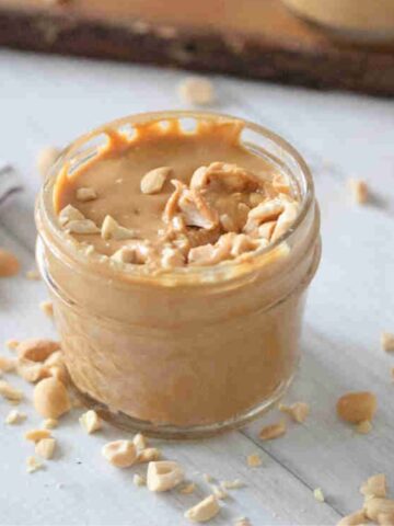 Homemade peanut butter in a mason jar topped with peanuts and more peanuts around it.