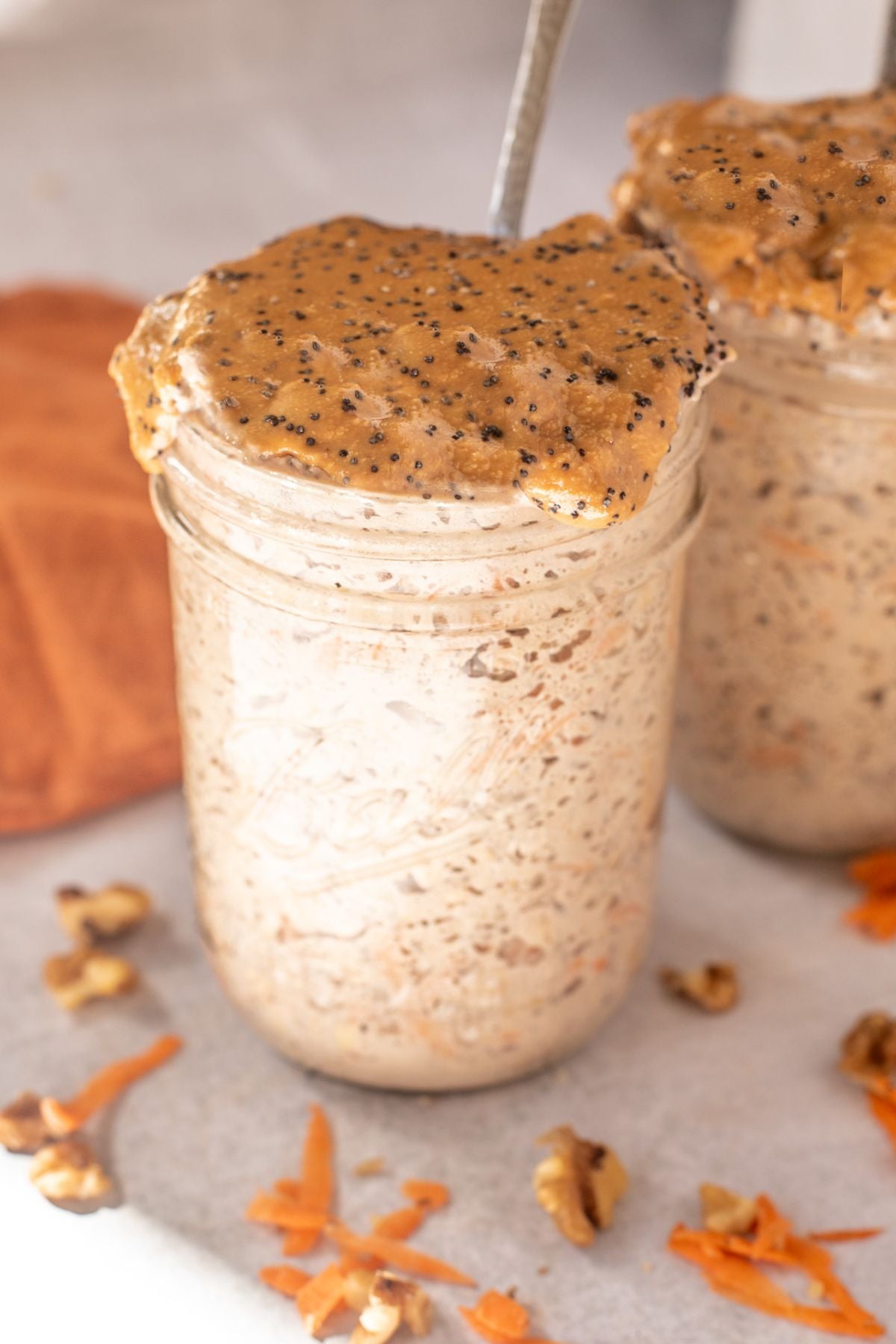 Two mason jars with carrot cake overnight oats topped with nut butter and chia seeds and a spoon in the first.