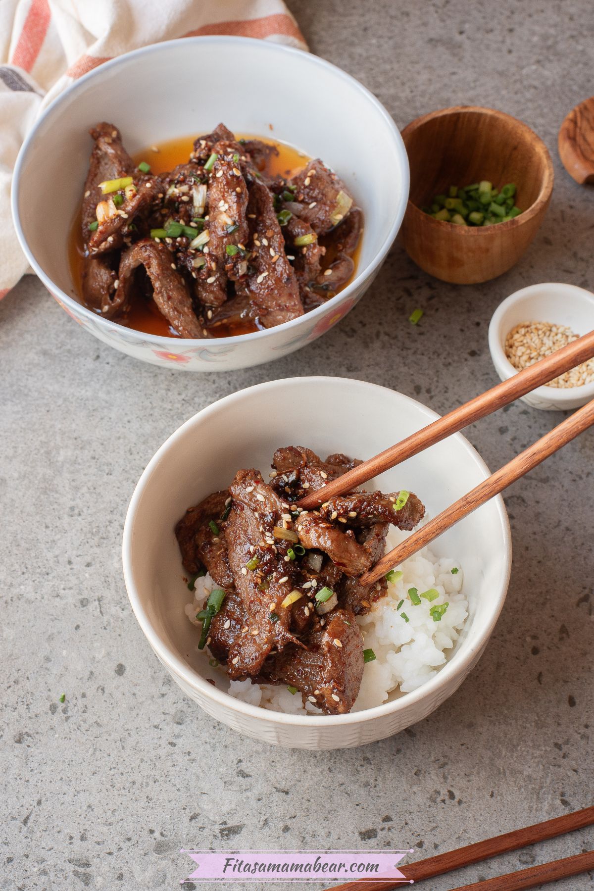A bowl of stir fried beef chili beside a bowl of rice with beef on top, and small bowls of sesame seeds and green onions beside them.