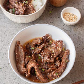 Two bowls of stir fried spicy beef, one topped on white rice, with a pair of chopsticks and small bowls of green onions and sesame seeds.