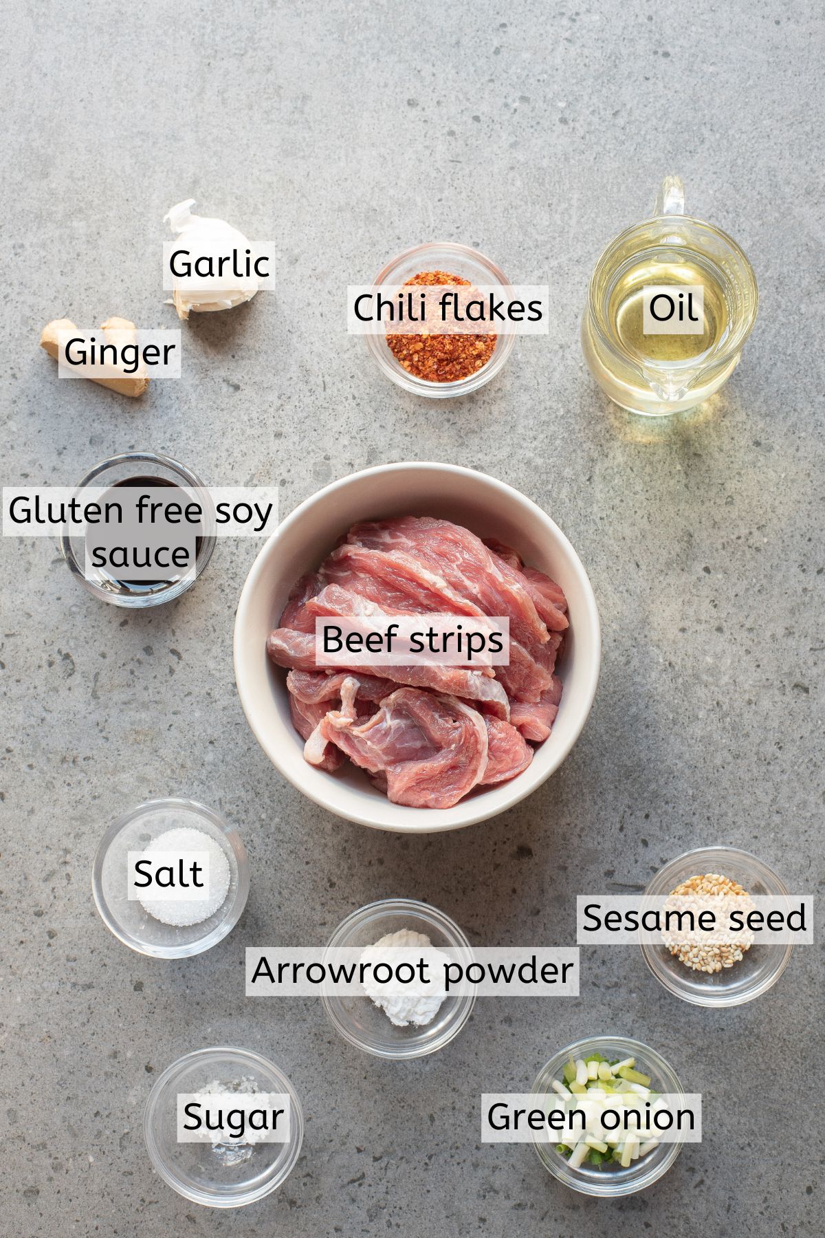 Ginger, garlic, oil, and bowls of soy sauce, beef strips, salt, arrowroot powder, sesame seed, sugar, and green onions.