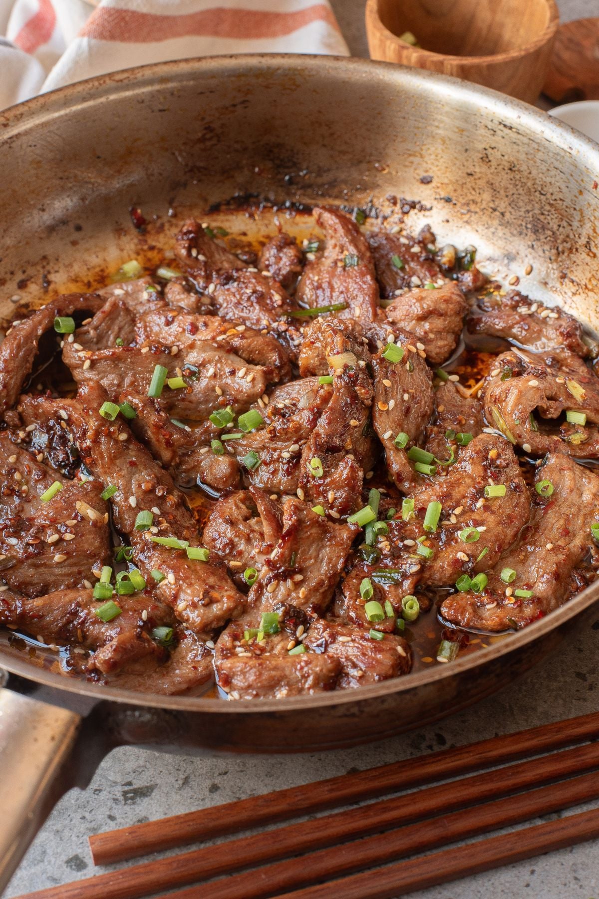 Seared and stir fried beef with chili in a pan with a small bowl of sesame seeds and green onions.