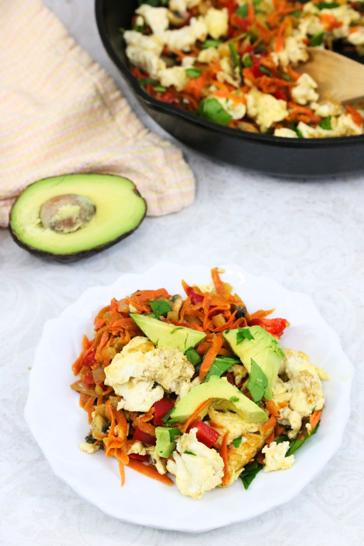 High protein egg scramble with avocados, carrots, and peppers, on a white plate with more in a skillet behind.