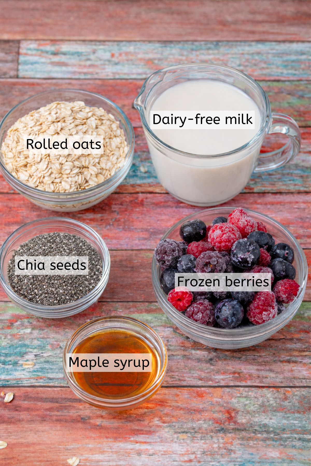 Dairy-free milk, berries, maple syrup, chia seeds, and oats in glass bowls with text labels over top.