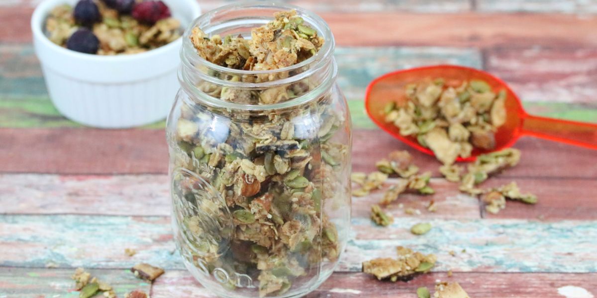 Homemade crunchy granola in a mason jar with more granola in a white ramekin and an orange scoop behind it.