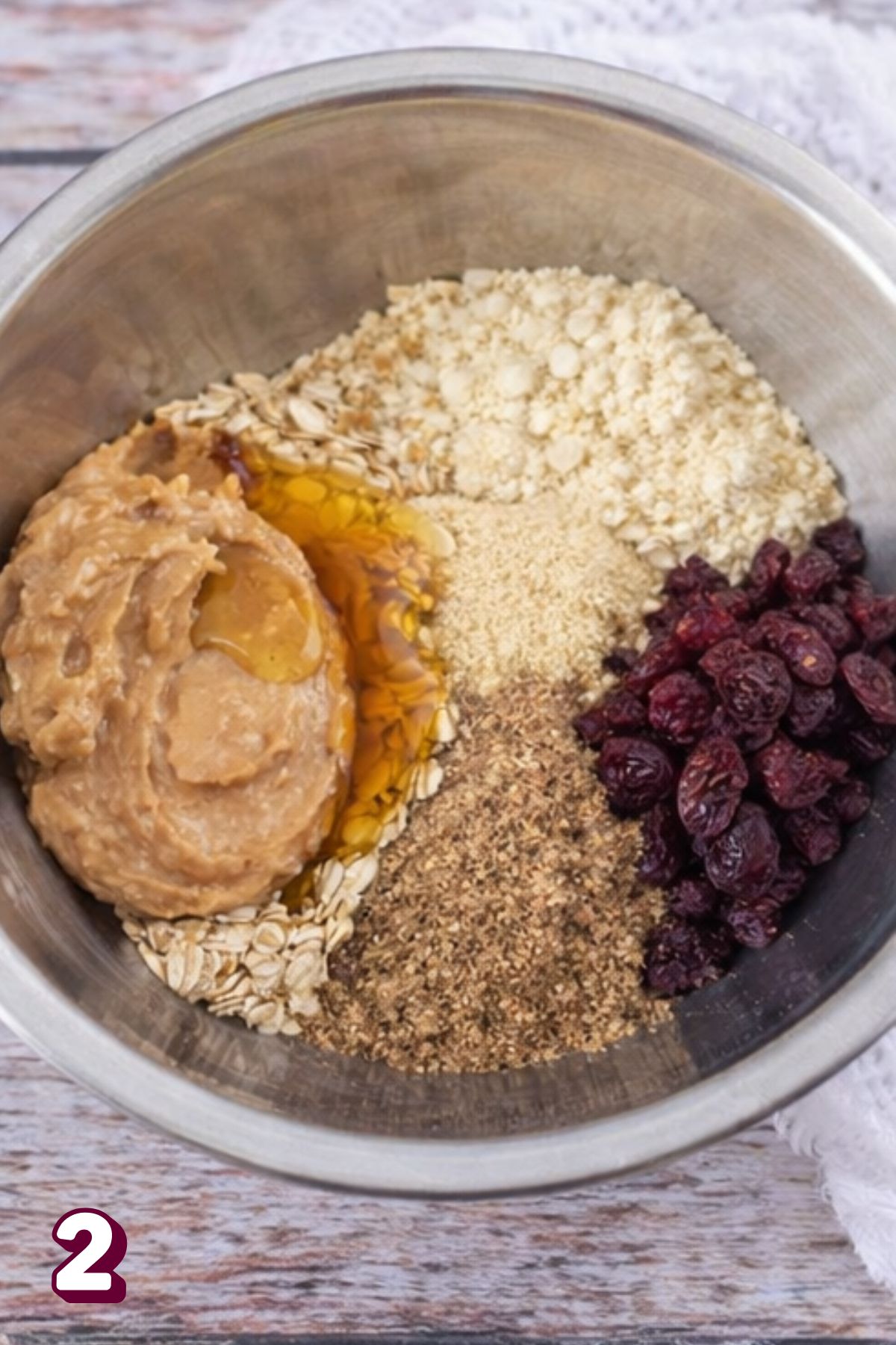 Steel bowl with flour, cranberries, nut butter, and oats in it with honey on top.