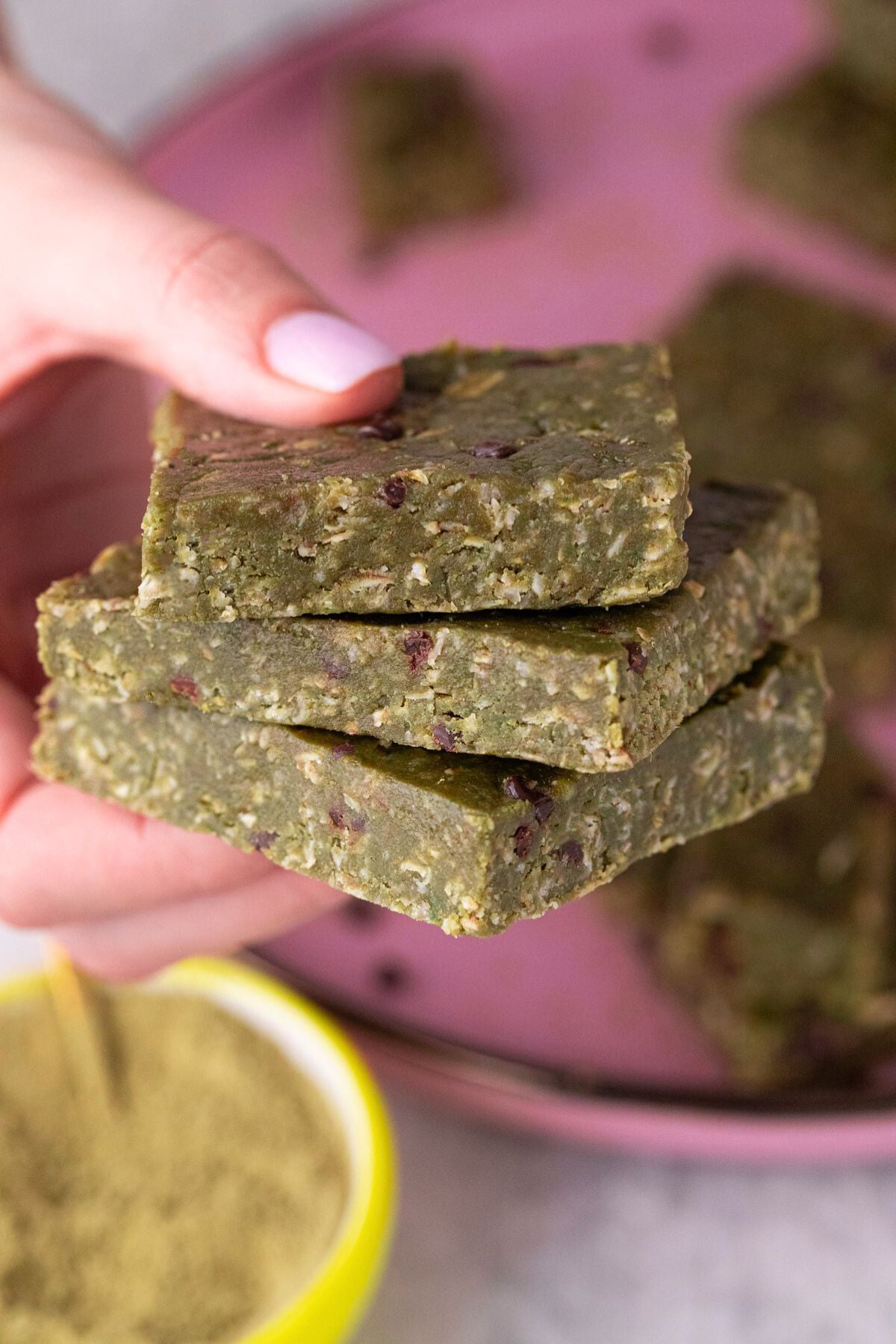Three matcha protein bars being held in fingers above a pink plate.