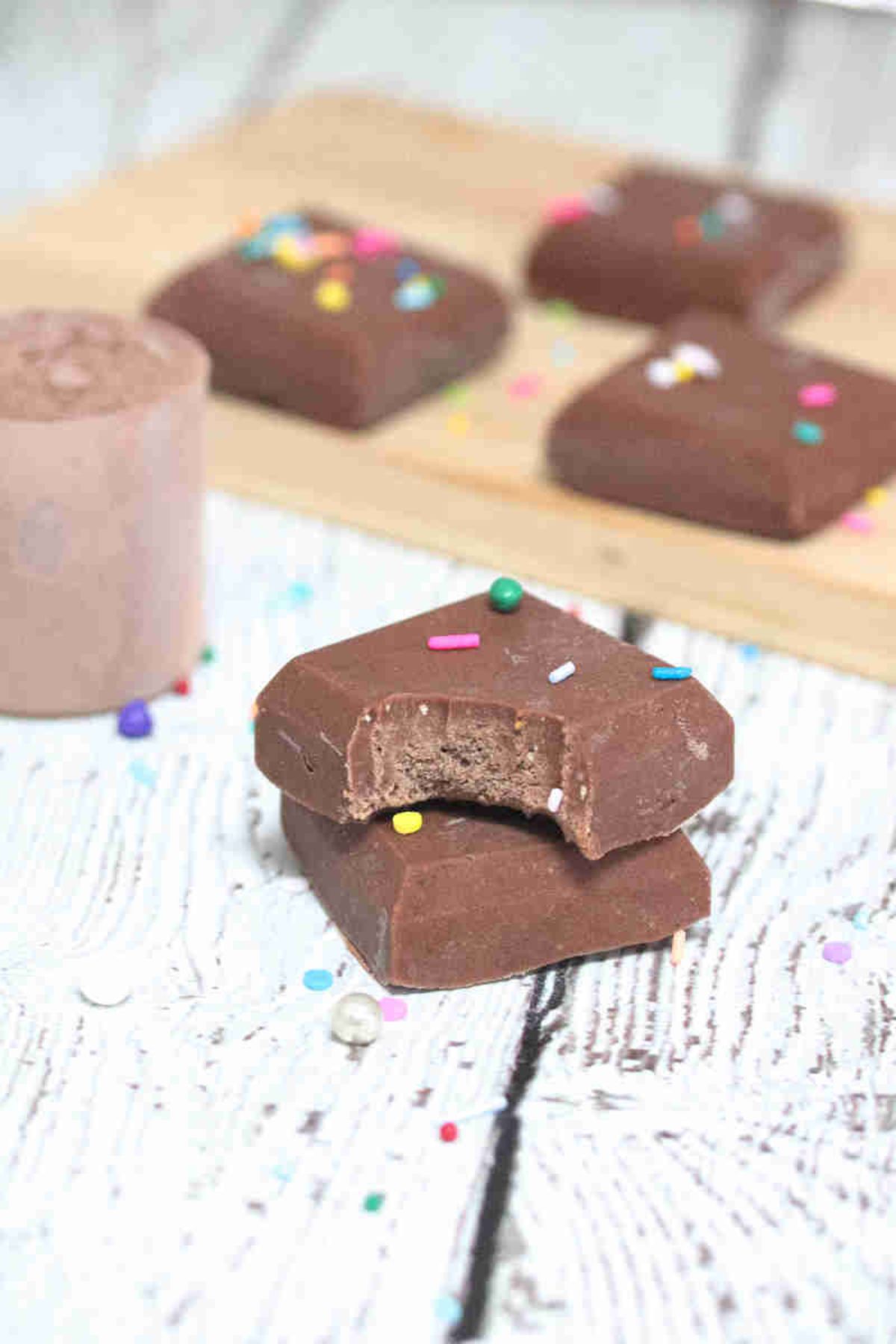 Protein powder chocolate fudge stacked up with chocolate chips on top and more fudge and protein powder behind it.