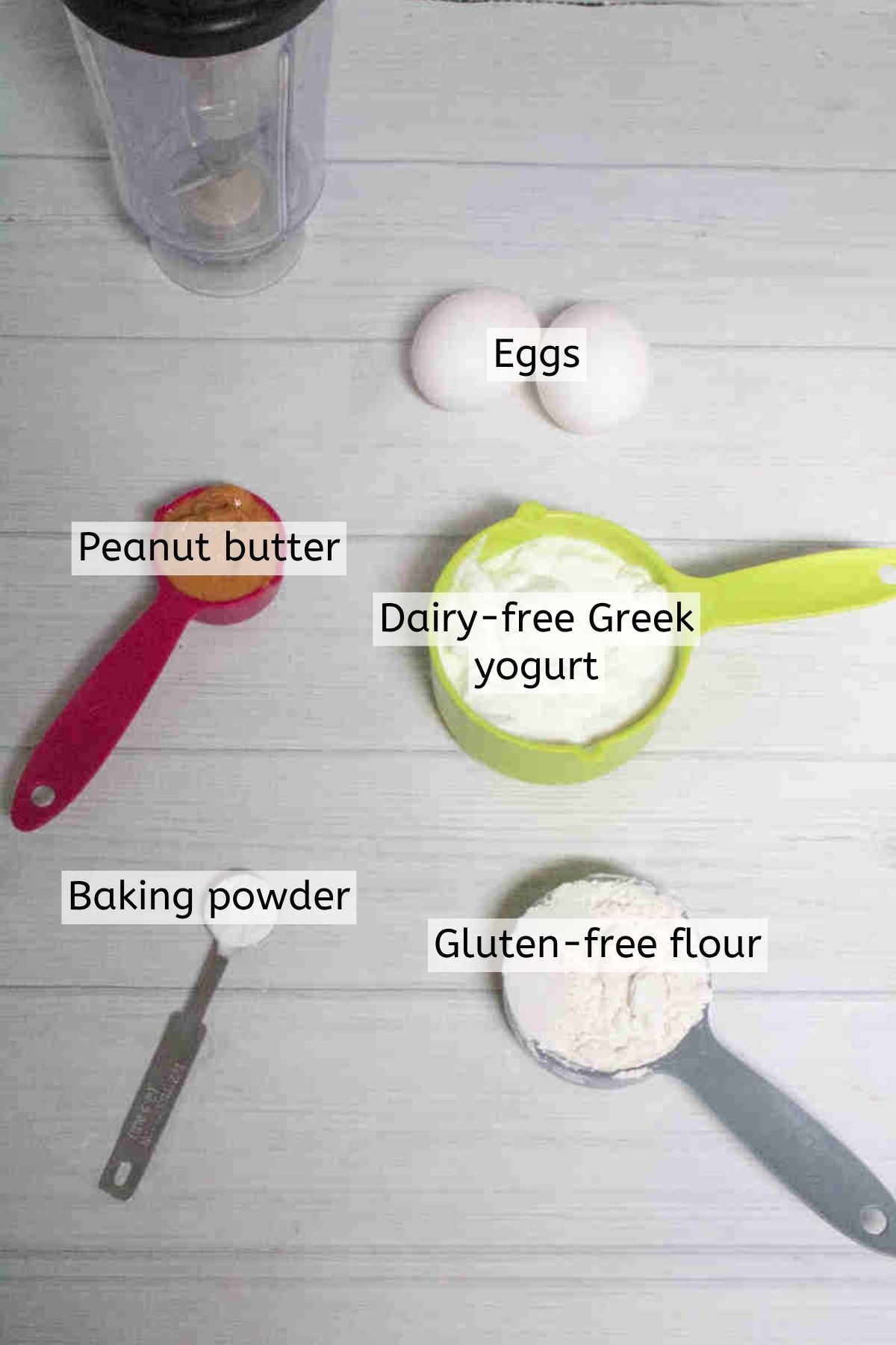 Greek yogurt, baking powder, peanut butter, and flour in measuring cups with text labels over top.