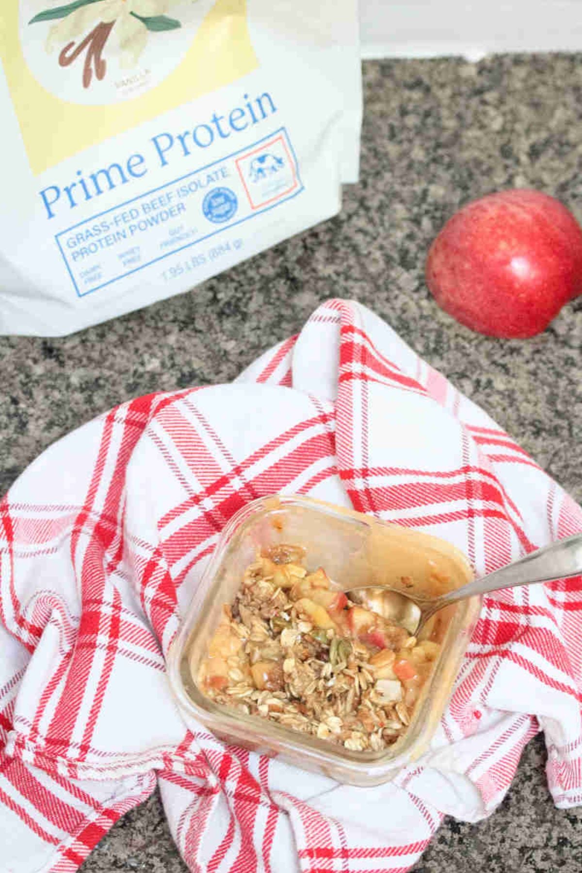 Protein apple crumble with oat topped in a single serve ramekin on a red linen with protein powder and an apple behind it.